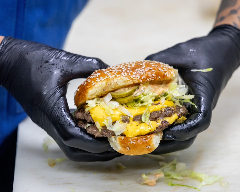 Gloved hands hold a double cheeseburger with lettuce, pickles, and special sauce on a sesame bun.