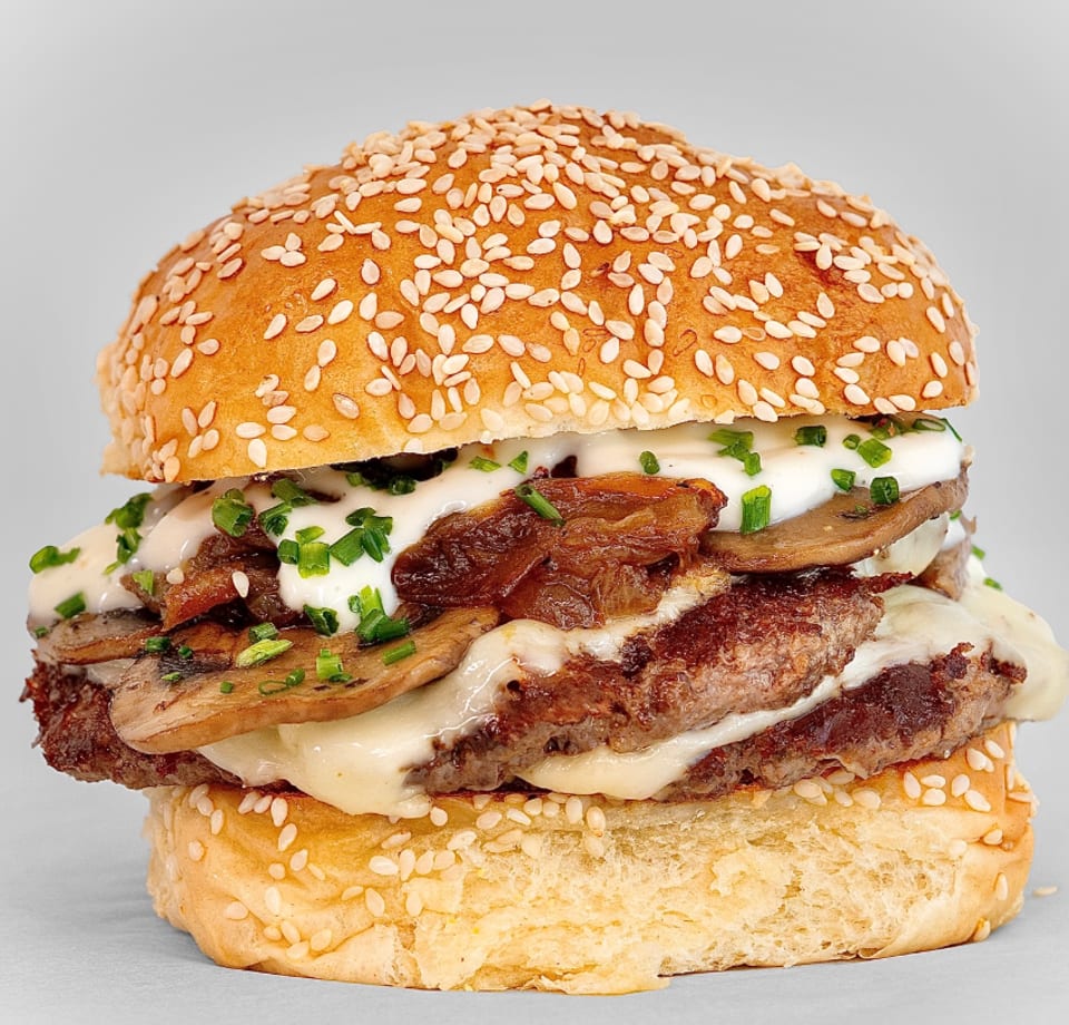Gourmet burger with two patties, melted cheese, mushrooms, caramelized onions, and chives on a sesame bun.