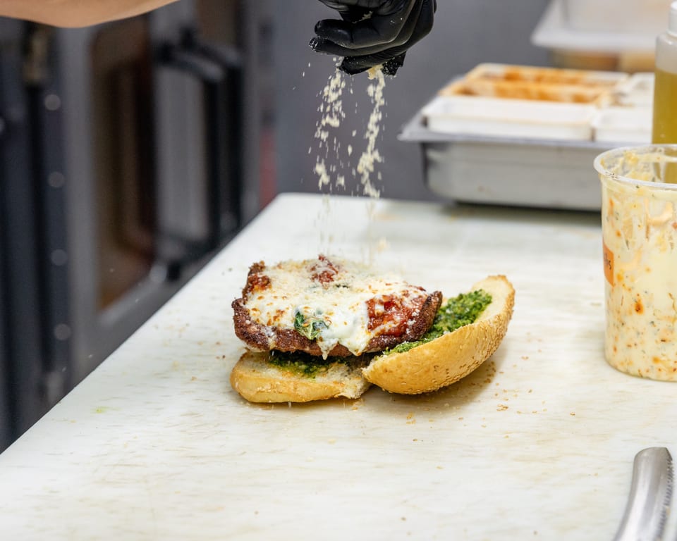 A chef's gloved hand sprinkles parmesan cheese onto a chicken parmesan sandwich with pesto.