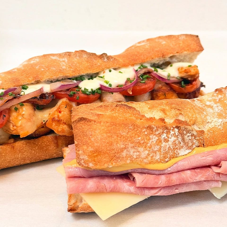 Two baguette sandwiches, one with chicken, tomatoes, and onions, the other with ham and cheese.
