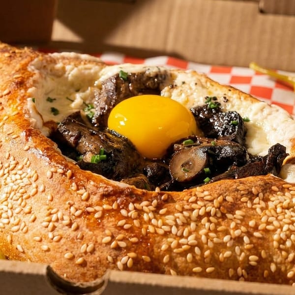 Khachapuri bread boat filled with mushrooms, cheese, and a raw egg yolk, in a takeout box.