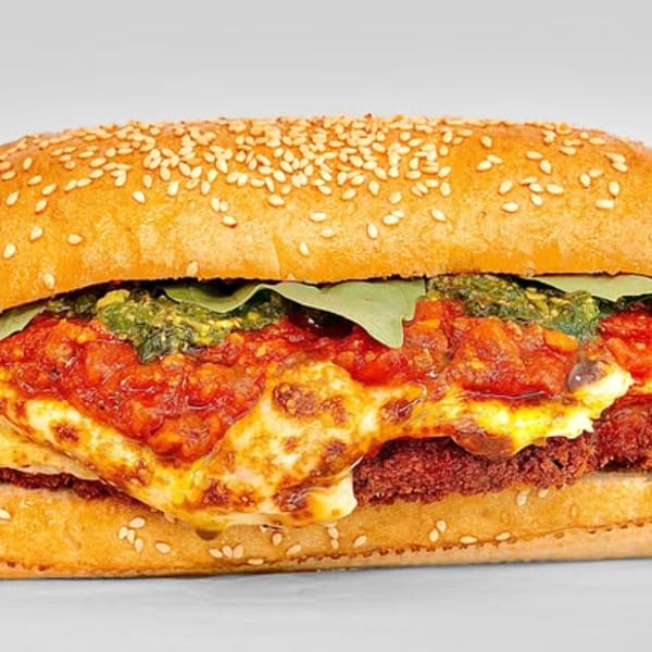 A close-up of a meatball sub with melted cheese, red sauce, and green pesto on a sesame seed bun.