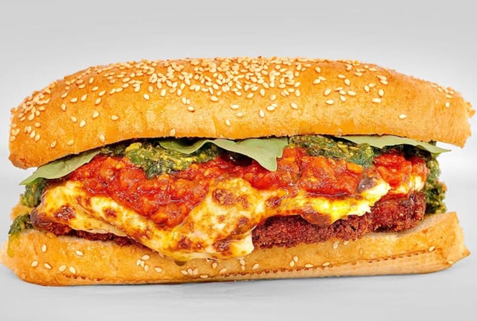 A close-up of a meatball sub with melted cheese, red sauce, and green pesto on a sesame seed bun.