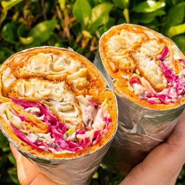 Two halves of a crispy chicken wrap with red cabbage and sauce, held against a green leafy background.