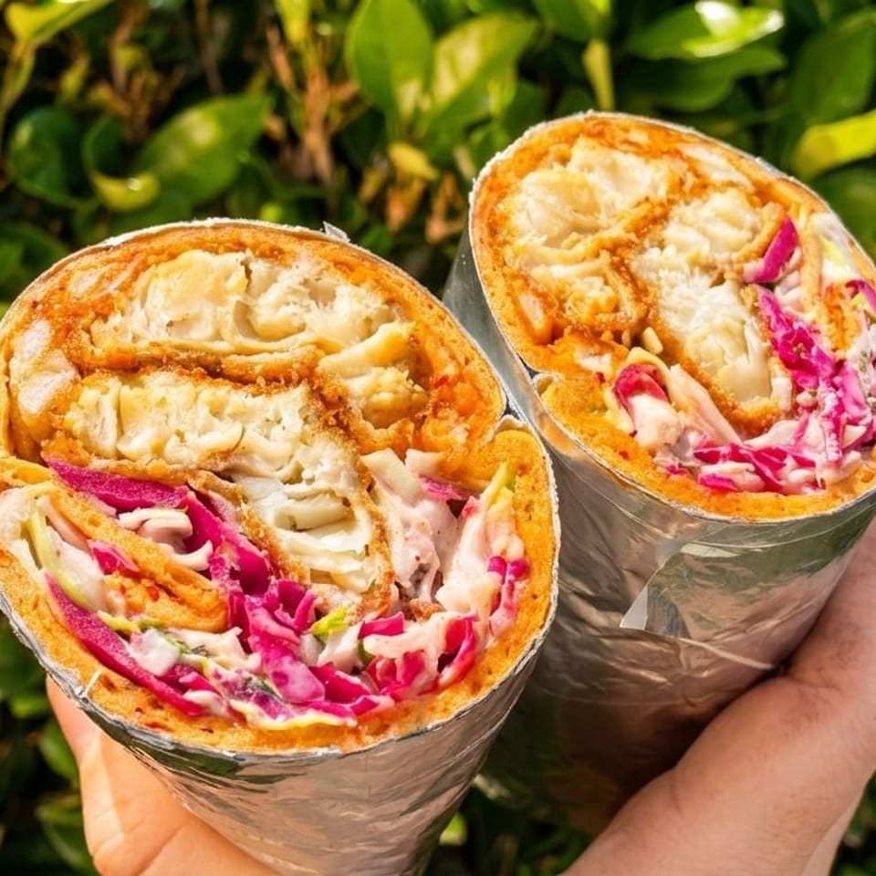 Two halves of a crispy chicken wrap with red cabbage and sauce, held against a green leafy background.