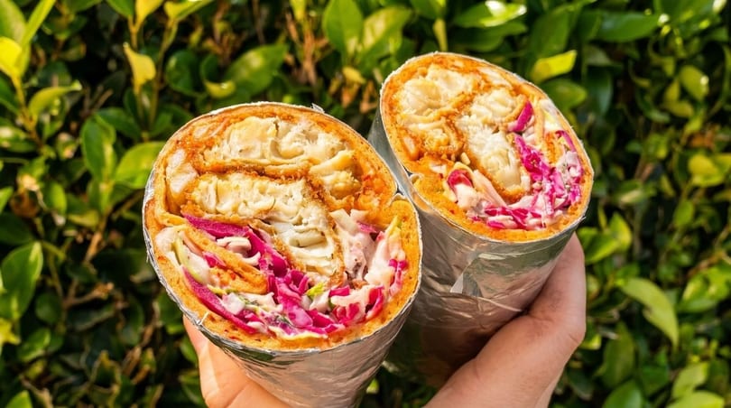 Two halves of a crispy chicken wrap with red cabbage and sauce, held against a green leafy background.